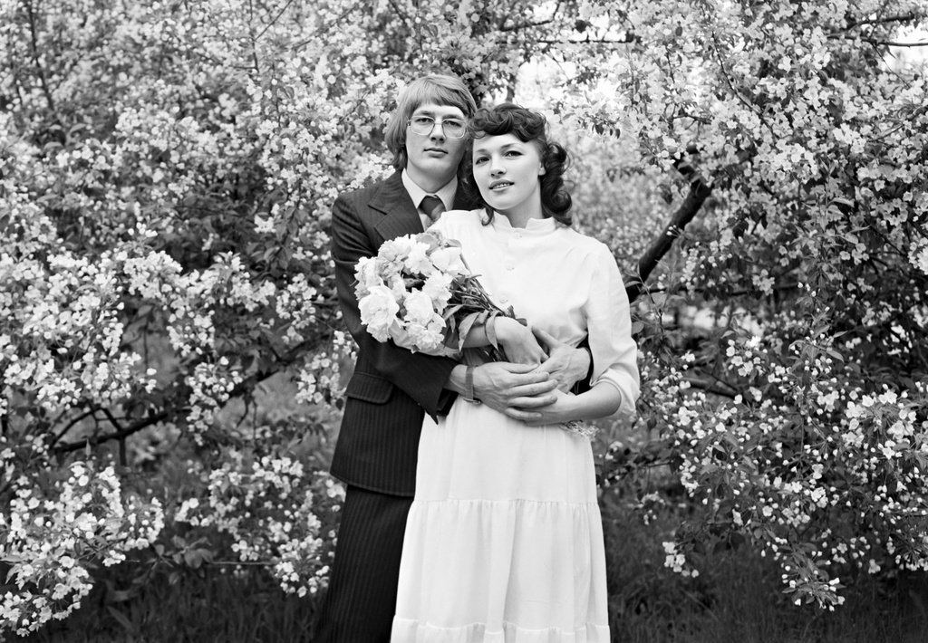Black-and-white wedding portrait of Lyudmila Chukhlomina and Anatoly Chukhlomin standing among blooming trees in the village of Siniy Utyos, Tomsk Region, on June 5, 1985.